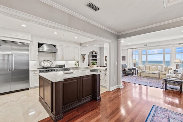 kitchen with wall chimney exhaust hood, dark brown cabinetry, ornate columns, ornamental molding, and stainless steel appliances