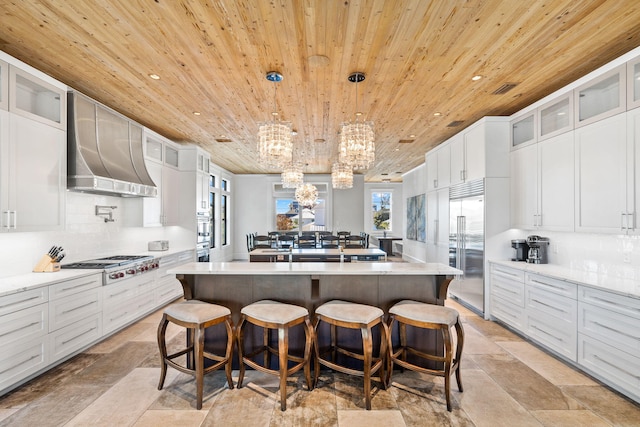 kitchen with wooden ceiling, wall chimney exhaust hood, an island with sink, appliances with stainless steel finishes, and decorative light fixtures