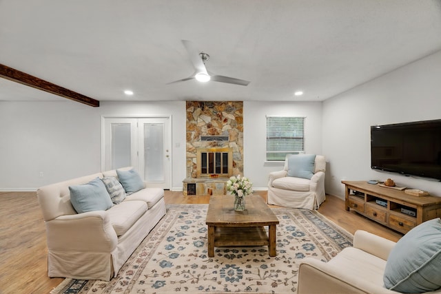 living room featuring beamed ceiling, light hardwood / wood-style floors, a stone fireplace, and ceiling fan