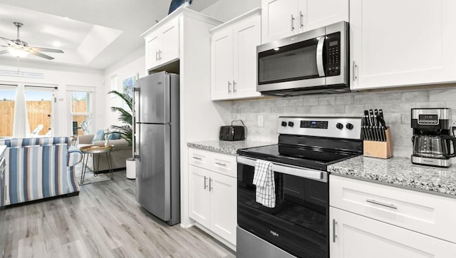 kitchen with ceiling fan, a raised ceiling, light stone countertops, stainless steel appliances, and white cabinets