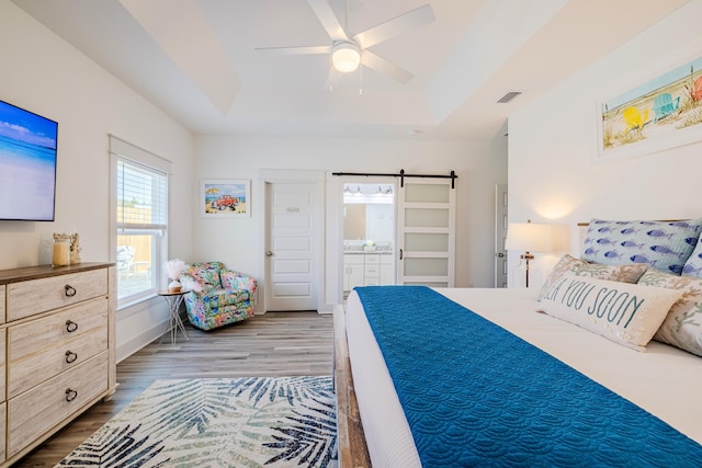 bedroom with ceiling fan, a barn door, a raised ceiling, hardwood / wood-style flooring, and connected bathroom