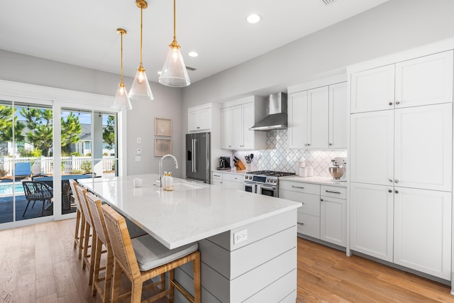 kitchen featuring a kitchen island with sink, wall chimney range hood, sink, appliances with stainless steel finishes, and white cabinetry