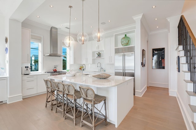 kitchen featuring built in fridge, white cabinets, an island with sink, and wall chimney exhaust hood