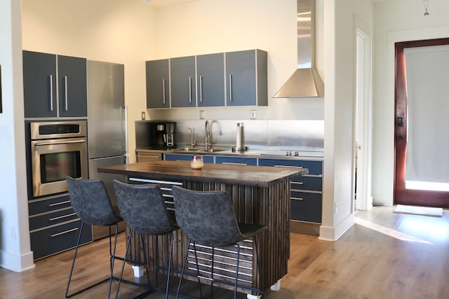 kitchen featuring sink, wall chimney range hood, butcher block countertops, a breakfast bar, and appliances with stainless steel finishes