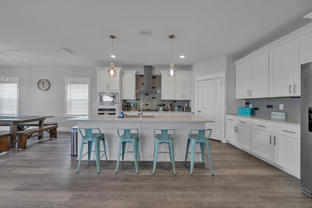 kitchen with white cabinets, hanging light fixtures, wall chimney exhaust hood, an island with sink, and stainless steel appliances