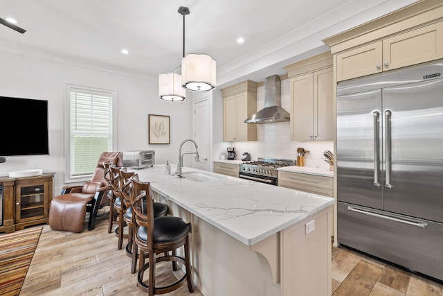 kitchen featuring wall chimney exhaust hood, sink, high end appliances, a center island with sink, and cream cabinets