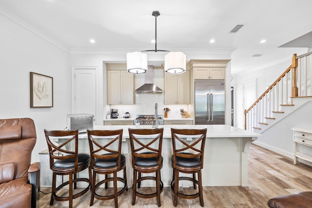 kitchen featuring built in fridge, an island with sink, hanging light fixtures, wall chimney exhaust hood, and cream cabinetry