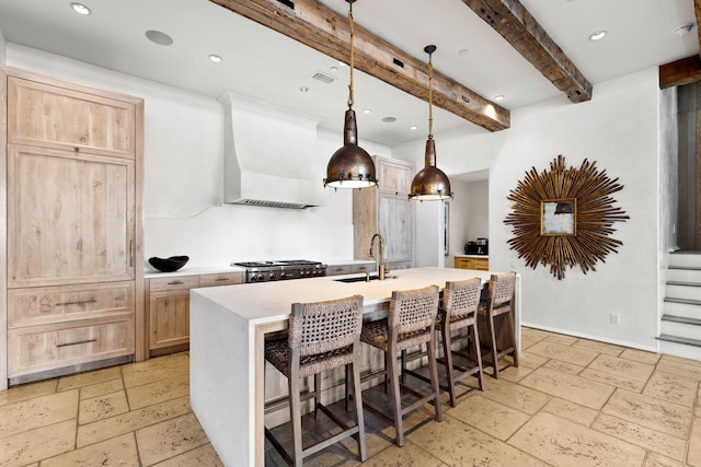 kitchen featuring light brown cabinets, wall chimney exhaust hood, hanging light fixtures, beamed ceiling, and a center island with sink