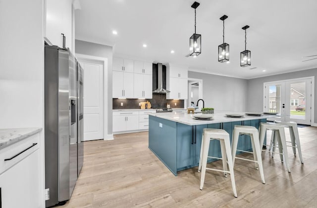 kitchen with a breakfast bar, a center island with sink, wall chimney range hood, stainless steel refrigerator with ice dispenser, and white cabinetry