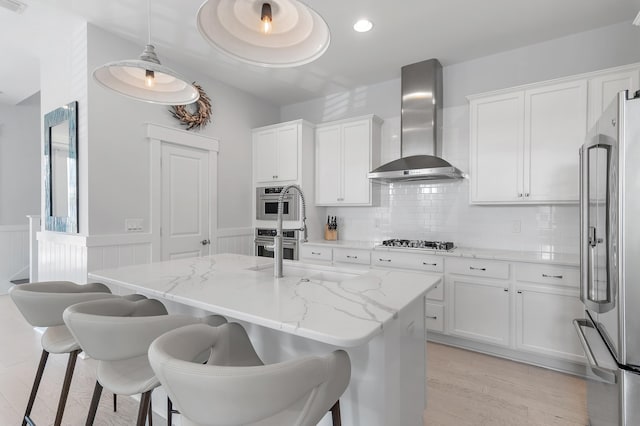 kitchen featuring a wainscoted wall, wall chimney exhaust hood, appliances with stainless steel finishes, and white cabinetry