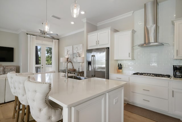 kitchen featuring appliances with stainless steel finishes, a kitchen island with sink, white cabinets, and wall chimney range hood