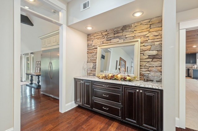 bar with light stone counters, dark hardwood / wood-style flooring, built in refrigerator, and dark brown cabinets