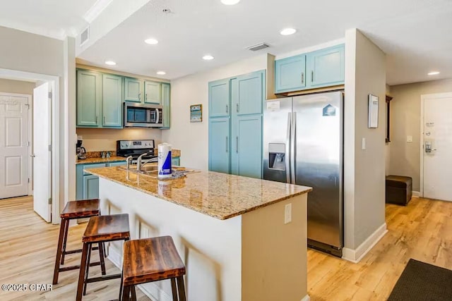 kitchen with light stone countertops, light hardwood / wood-style flooring, stainless steel appliances, a kitchen island with sink, and a kitchen breakfast bar