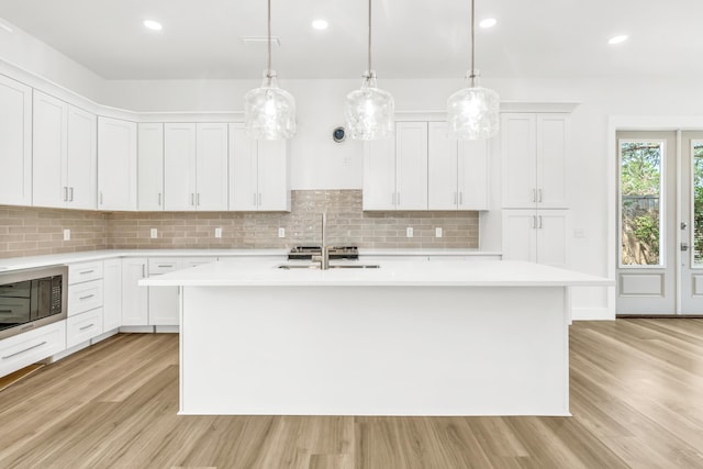 kitchen featuring a kitchen island with sink, hanging light fixtures, black microwave, decorative backsplash, and white cabinetry