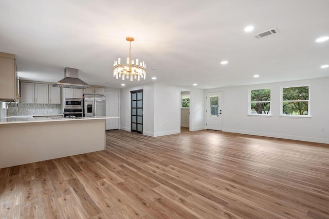 kitchen featuring stainless steel appliances, light hardwood / wood-style flooring, decorative backsplash, hanging light fixtures, and wall chimney range hood