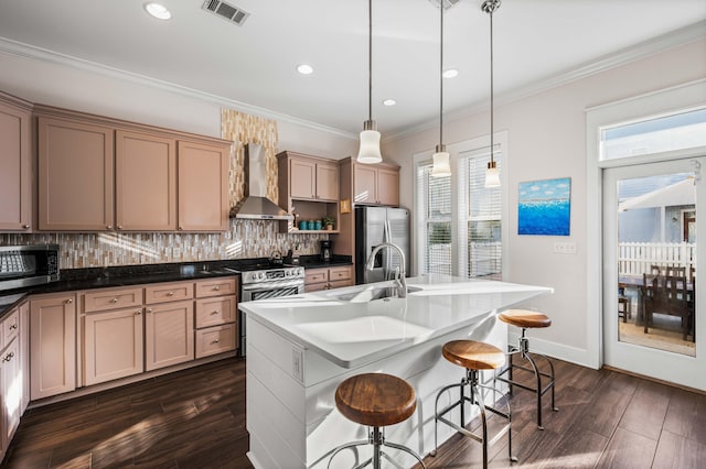 kitchen featuring wall chimney range hood, a kitchen island with sink, hanging light fixtures, stainless steel appliances, and a kitchen bar