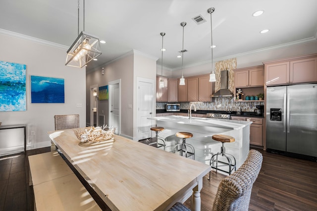 kitchen with appliances with stainless steel finishes, decorative light fixtures, an island with sink, backsplash, and wall chimney range hood