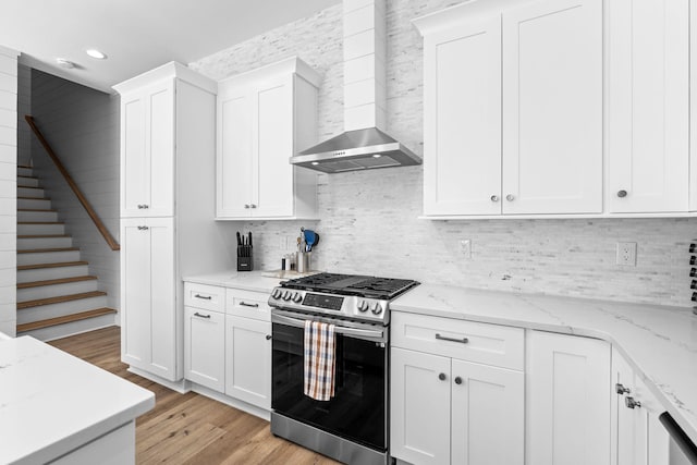 kitchen with light stone counters, light hardwood / wood-style flooring, wall chimney range hood, stainless steel gas stove, and white cabinets