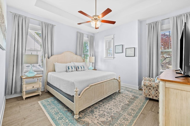 bedroom featuring multiple windows, ceiling fan, a tray ceiling, and light wood-type flooring