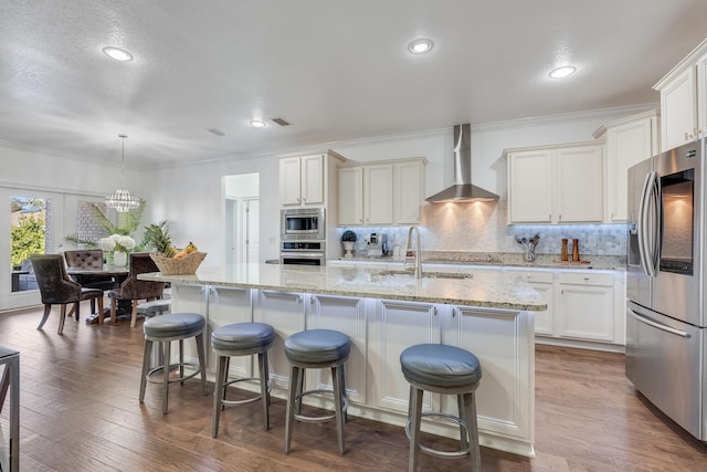 kitchen featuring wall chimney range hood, pendant lighting, a center island with sink, sink, and appliances with stainless steel finishes