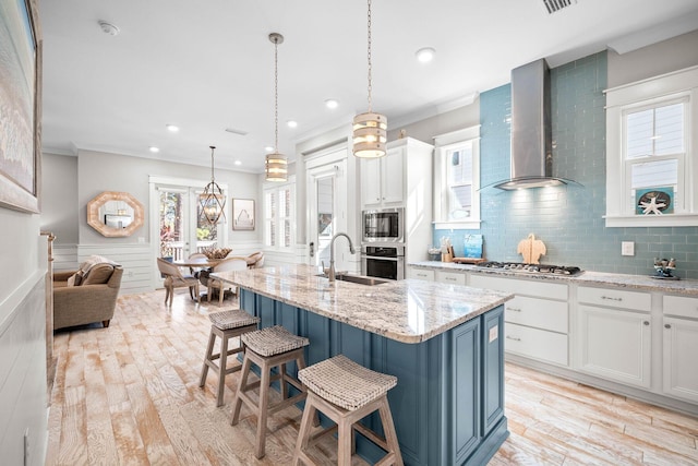 kitchen featuring wall chimney exhaust hood, a kitchen island with sink, white cabinets, appliances with stainless steel finishes, and sink