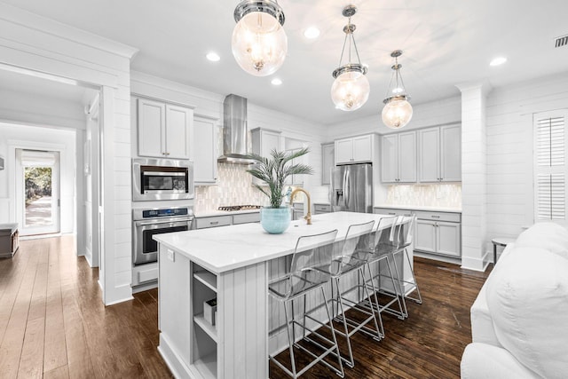 kitchen featuring a breakfast bar, pendant lighting, wall chimney range hood, stainless steel appliances, and a center island with sink