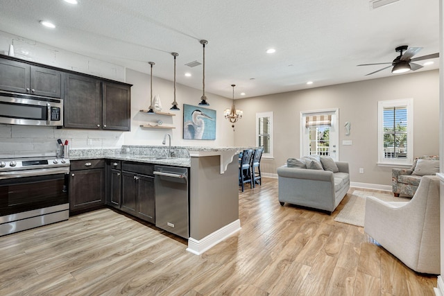 kitchen featuring pendant lighting, light hardwood / wood-style floors, appliances with stainless steel finishes, light stone counters, and ceiling fan with notable chandelier