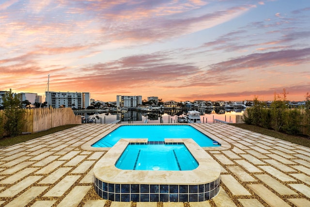 pool at dusk with a water view, a patio area, and an in ground hot tub