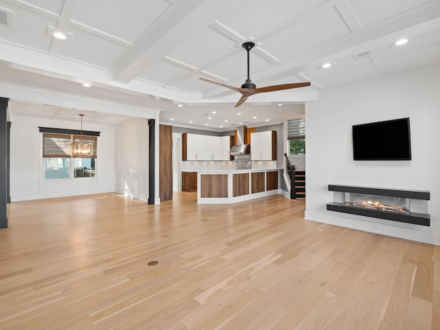 unfurnished living room featuring ceiling fan with notable chandelier, a wainscoted wall, light wood finished floors, beamed ceiling, and a glass covered fireplace