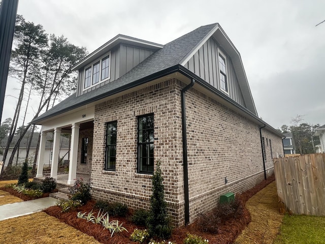 view of property exterior featuring a shingled roof, covered porch, fence, board and batten siding, and brick siding