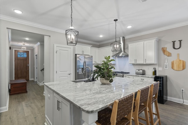 kitchen featuring wall chimney range hood, sink, stainless steel fridge, white cabinetry, and an island with sink