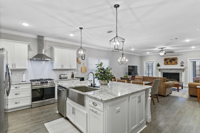 kitchen with appliances with stainless steel finishes, decorative light fixtures, white cabinetry, a kitchen island with sink, and wall chimney exhaust hood