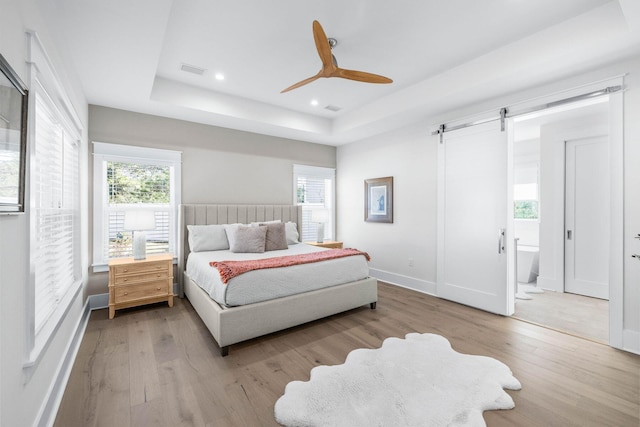 bedroom featuring a raised ceiling, a barn door, ceiling fan, and light hardwood / wood-style flooring