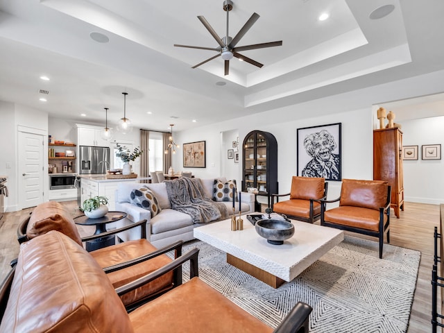 living room with ceiling fan, a raised ceiling, and light wood-type flooring