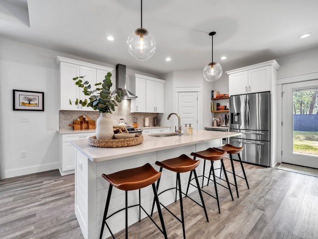 kitchen featuring pendant lighting, white cabinets, stainless steel fridge, a kitchen island with sink, and wall chimney exhaust hood