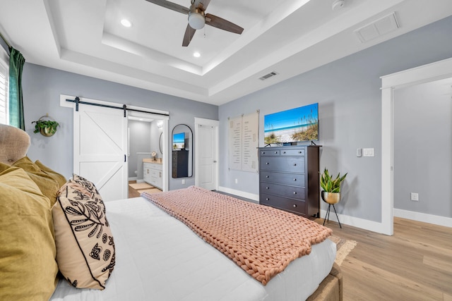 bedroom featuring a barn door, light wood-type flooring, a raised ceiling, ceiling fan, and connected bathroom