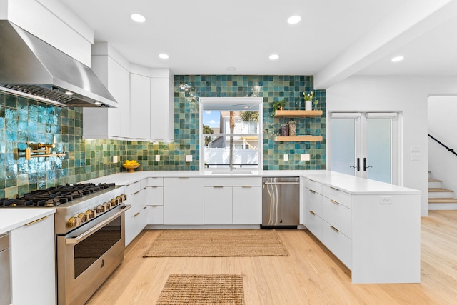 kitchen with wall chimney exhaust hood, white cabinetry, stainless steel appliances, tasteful backsplash, and light hardwood / wood-style flooring