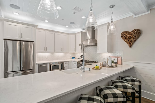 kitchen featuring appliances with stainless steel finishes, decorative light fixtures, white cabinetry, wall chimney range hood, and a breakfast bar area