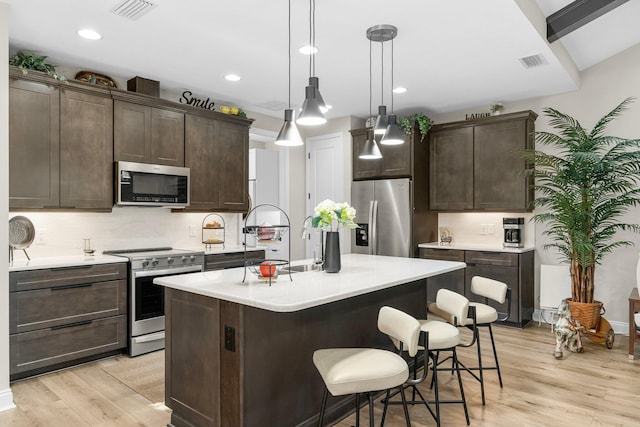 kitchen featuring appliances with stainless steel finishes, dark brown cabinetry, light hardwood / wood-style floors, an island with sink, and decorative light fixtures