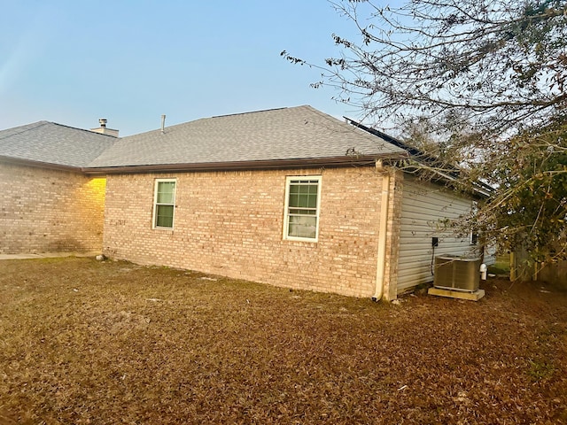 view of side of property with central air condition unit, brick siding, a lawn, and roof with shingles