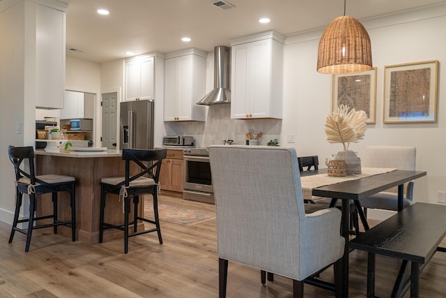 kitchen with pendant lighting, wall chimney range hood, appliances with stainless steel finishes, white cabinetry, and light wood-type flooring