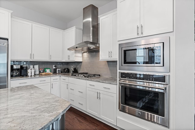 kitchen with white cabinetry, decorative backsplash, light stone counters, stainless steel appliances, and wall chimney range hood