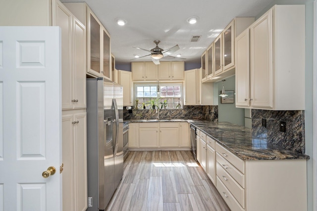 kitchen featuring backsplash, glass insert cabinets, appliances with stainless steel finishes, a ceiling fan, and dark stone counters