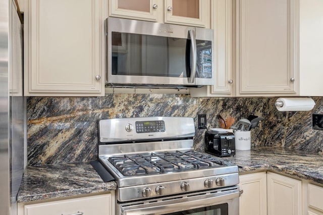 kitchen with cream cabinets, appliances with stainless steel finishes, dark stone counters, tasteful backsplash, and glass insert cabinets