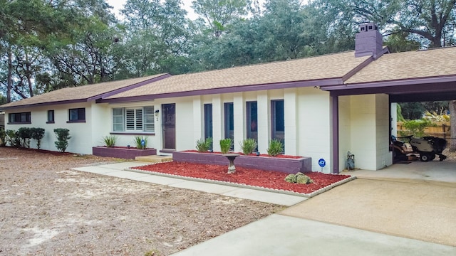 single story home featuring an attached carport, a shingled roof, concrete block siding, concrete driveway, and a chimney