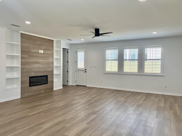 unfurnished living room featuring a large fireplace, built in shelves, ceiling fan, and light hardwood / wood-style flooring