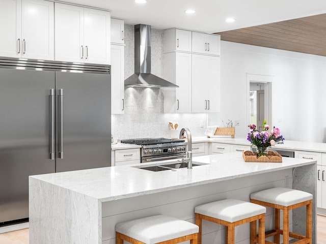 kitchen with an island with sink, white cabinets, high end appliances, wall chimney range hood, and a breakfast bar area