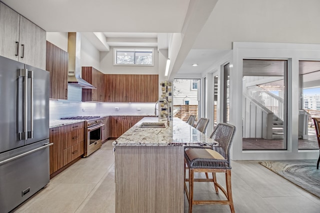 kitchen featuring a breakfast bar, high quality appliances, sink, a kitchen island with sink, and wall chimney range hood