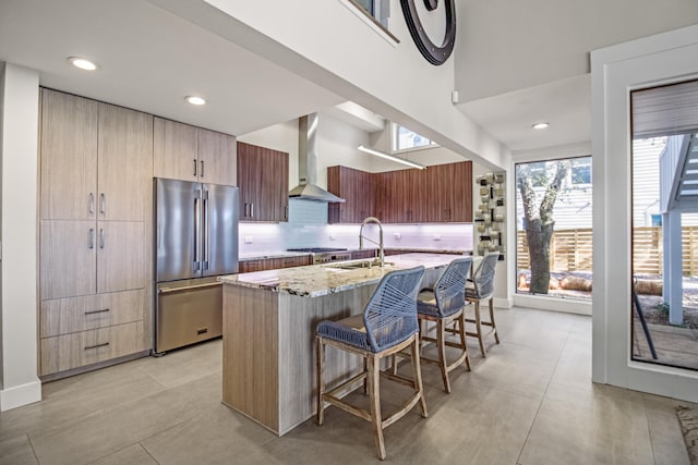 kitchen with wall chimney range hood, appliances with stainless steel finishes, a kitchen breakfast bar, light stone counters, and a center island with sink