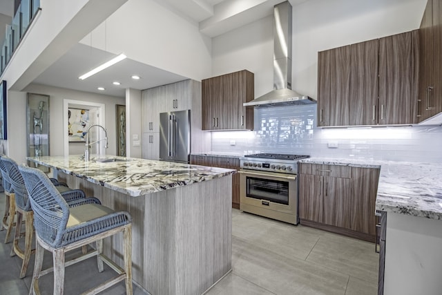 kitchen with wall chimney exhaust hood, sink, light stone counters, premium appliances, and a kitchen island with sink